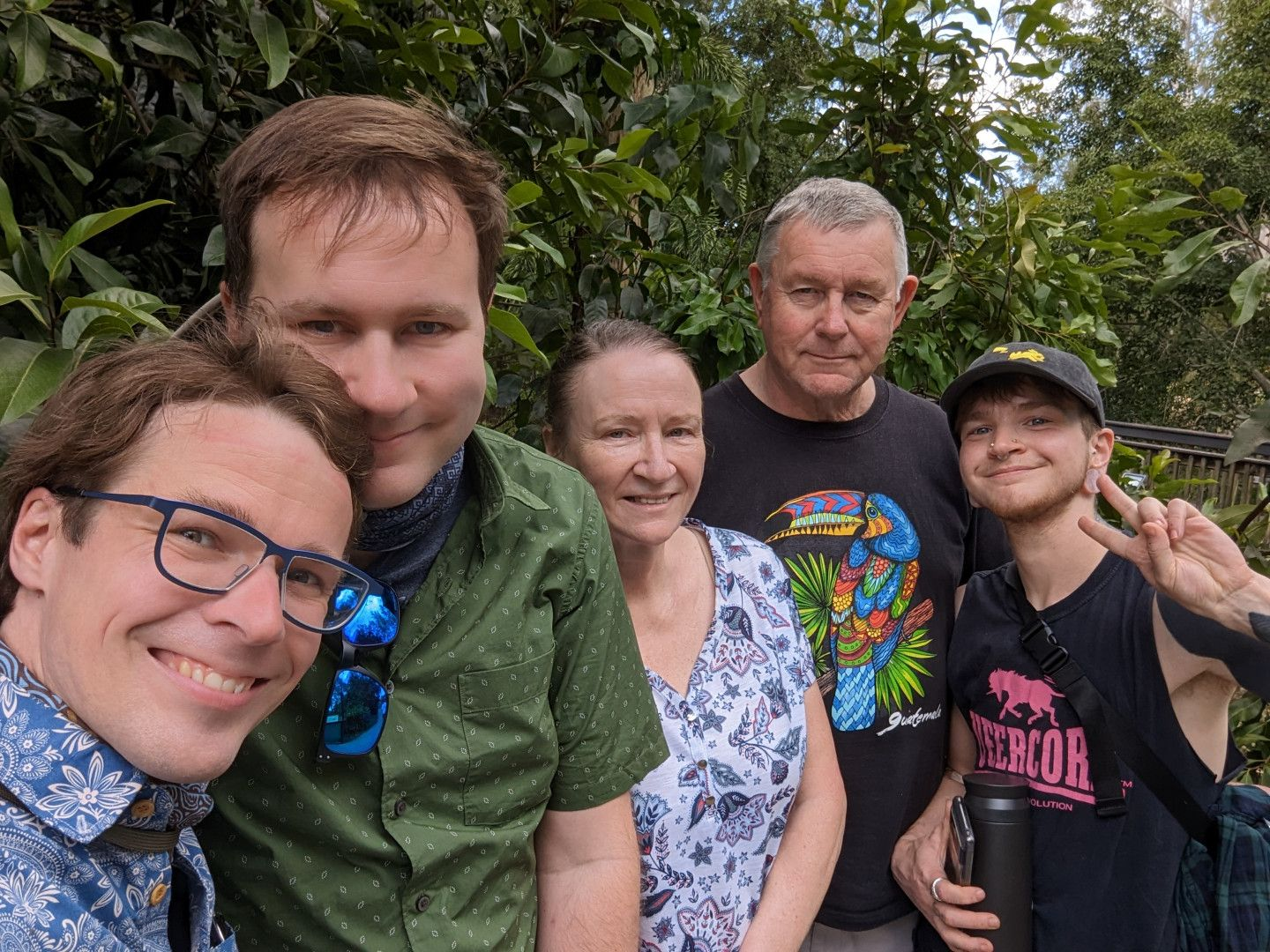 A photo of the whole Aussie family that Josh married into, from left to right: Josh, Chris, Michelle, Rolf, and Rory. Everyone is smiling, to some degree, and Rory is throwing a peace town. Behind them is a stand of verdant subtropical plants