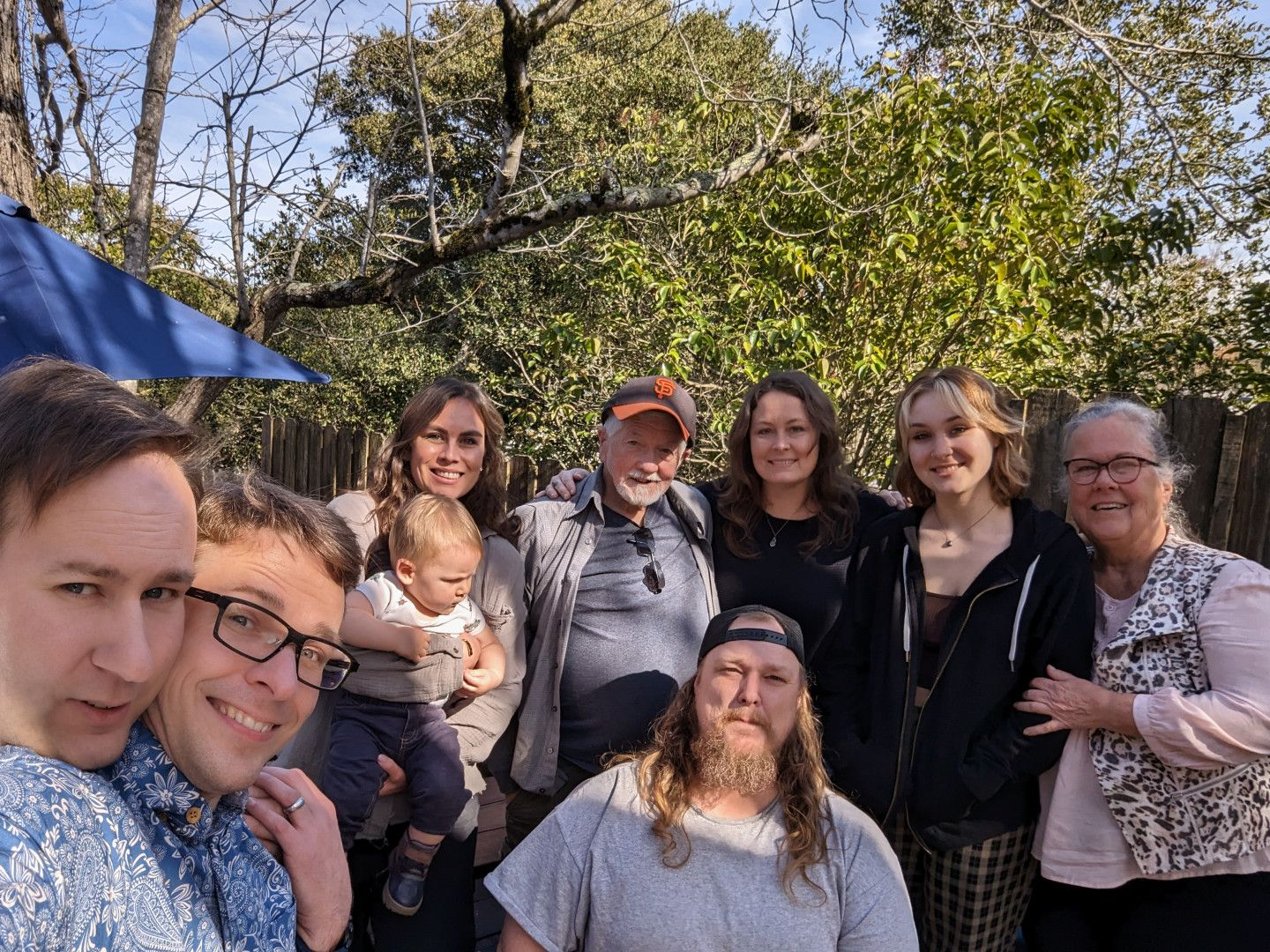 A family photo taken on a sunny winter day in our backyard. Lots of smiling faces against a backdrop of happy trees. From left to right: Chris, Josh, Jen holding Hunter, Carl, Greg in front, Abra, Shasta, and Donna.