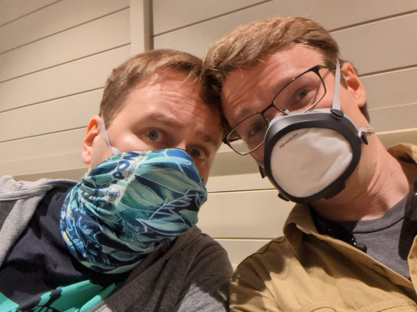 A photo of Josh and Chris leaning their heads together looking a mix of happy and concerned. They are indoors at the airport, wearing elastomeric masks.